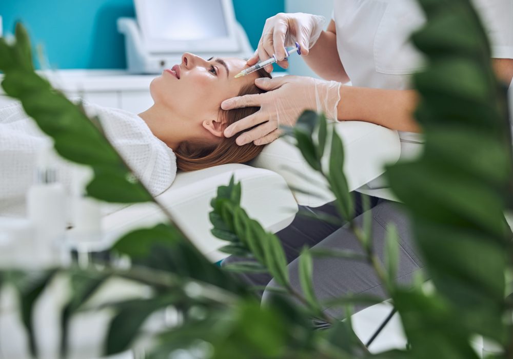 Waist up side view portrait of elegant redheaded woman lying on the medicine chair while beautician taking face injection with special product in beauty center